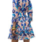 A woman is seen from the back wearing the Figue Cami Dress, which features a ruffled, long-sleeve design with an exaggerated shirt collar and a blue, pink, and beige abstract pattern.