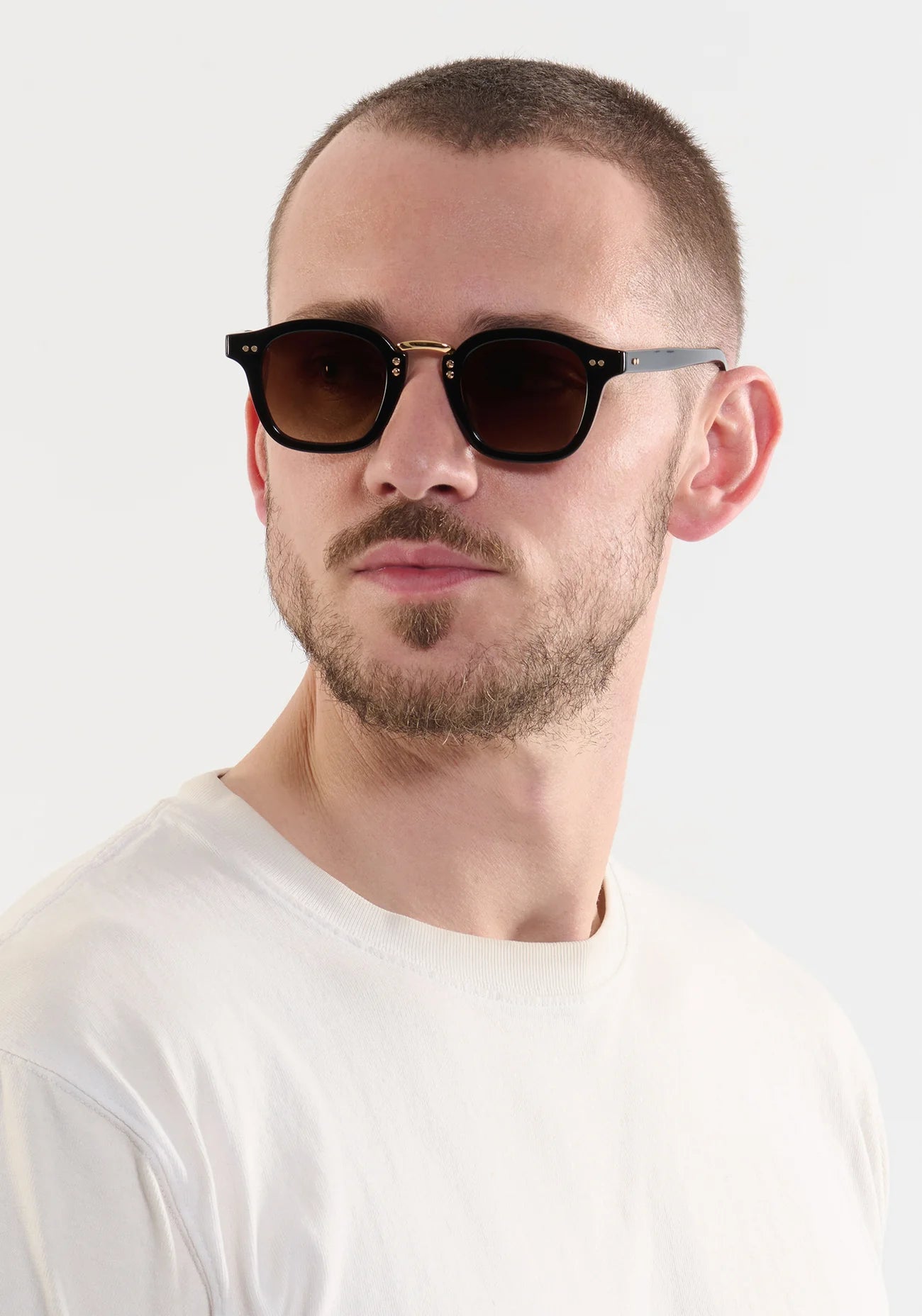 A man with short hair and a trimmed beard wears Krewe Campbell Sunglasses—round, black, hand-crafted acetate—and a plain white t-shirt, gazing to the side against a light background.