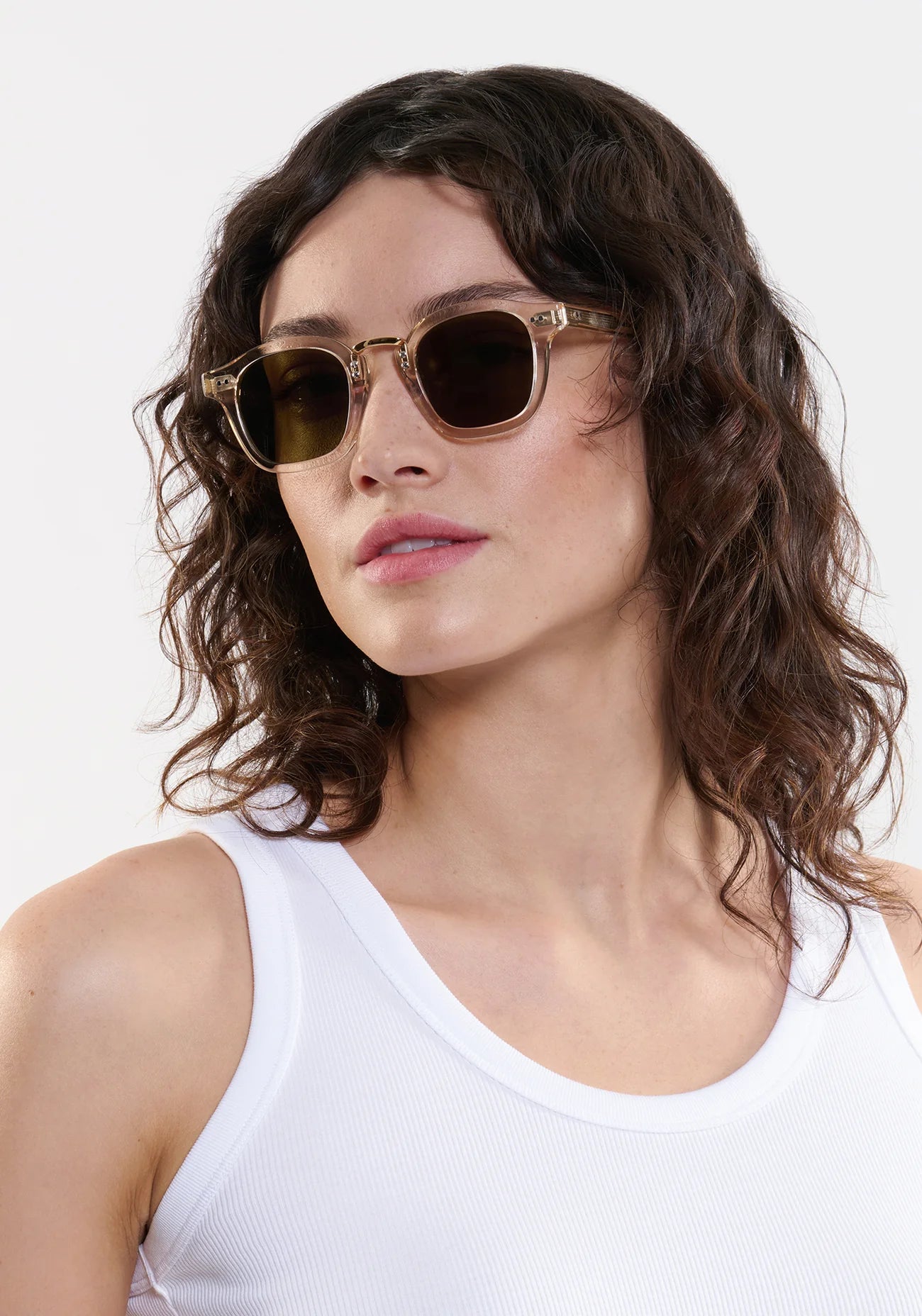 A woman with wavy brown hair wears Krewe's Campbell Polarized Sunglasses in brown acetate and a white sleeveless top, gazing slightly to the side against a plain background.