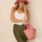 A woman wears the Lorna Murray Island Capri Hat—a wide-brimmed pink straw sun hat with UPF 50+ protection—paired with a white sleeveless top, olive skirt, and a matching pink straw tote bag against a beige background, creating the perfect look for stylish travel or sunny adventures.