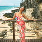 A woman in a patterned Paloma Wrap Shirt by Hunter Bell NYC stands on a wooden deck with her eyes closed and arms slightly outstretched, against a backdrop of rocky beach and ocean.