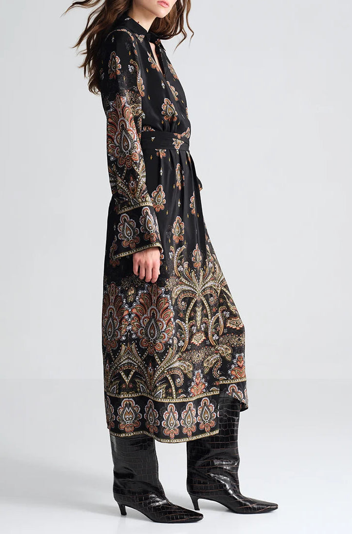 A woman wears the Viv Midi Dress by Cara Cara—black, faux wrap, paisley-print with long sleeves and belt—plus black knee-high boots, against a plain light background.