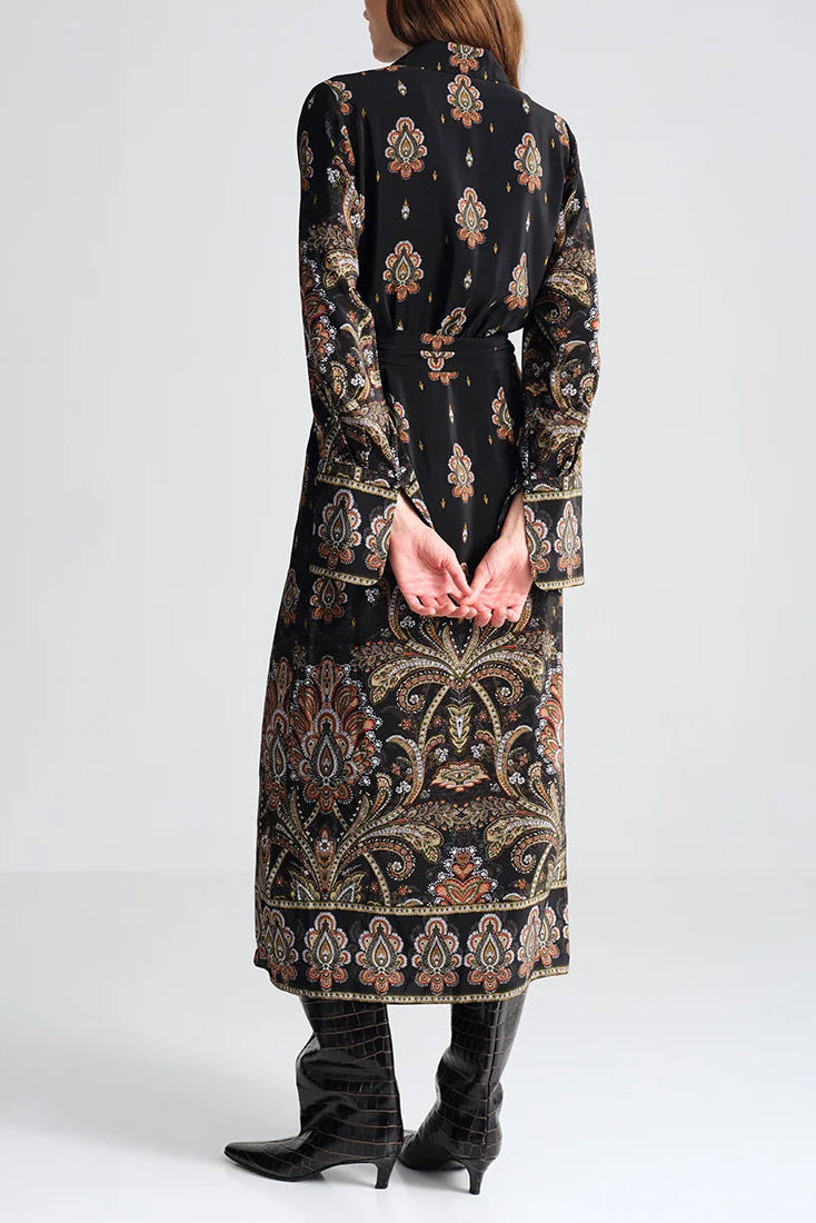 Person stands with hands behind back, wearing the Cara Cara Viv Midi Dress—long sleeve, black with paisley patterns—and black boots, facing away from the camera against a plain background.