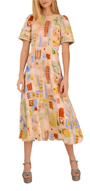 A woman models the Casey Dress by Hunter Bell NYC—a flattering, pastel sheath midi dress with abstract patterns and short sleeves—paired with silver platform sandals against a white background.