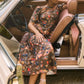 A woman wearing the Casey Dress by Hunter Bell NYC, a universally flattering floral sheath midi, sits in the driver’s seat of a vintage beige convertible with the door open, her arm resting on the back of the seat.