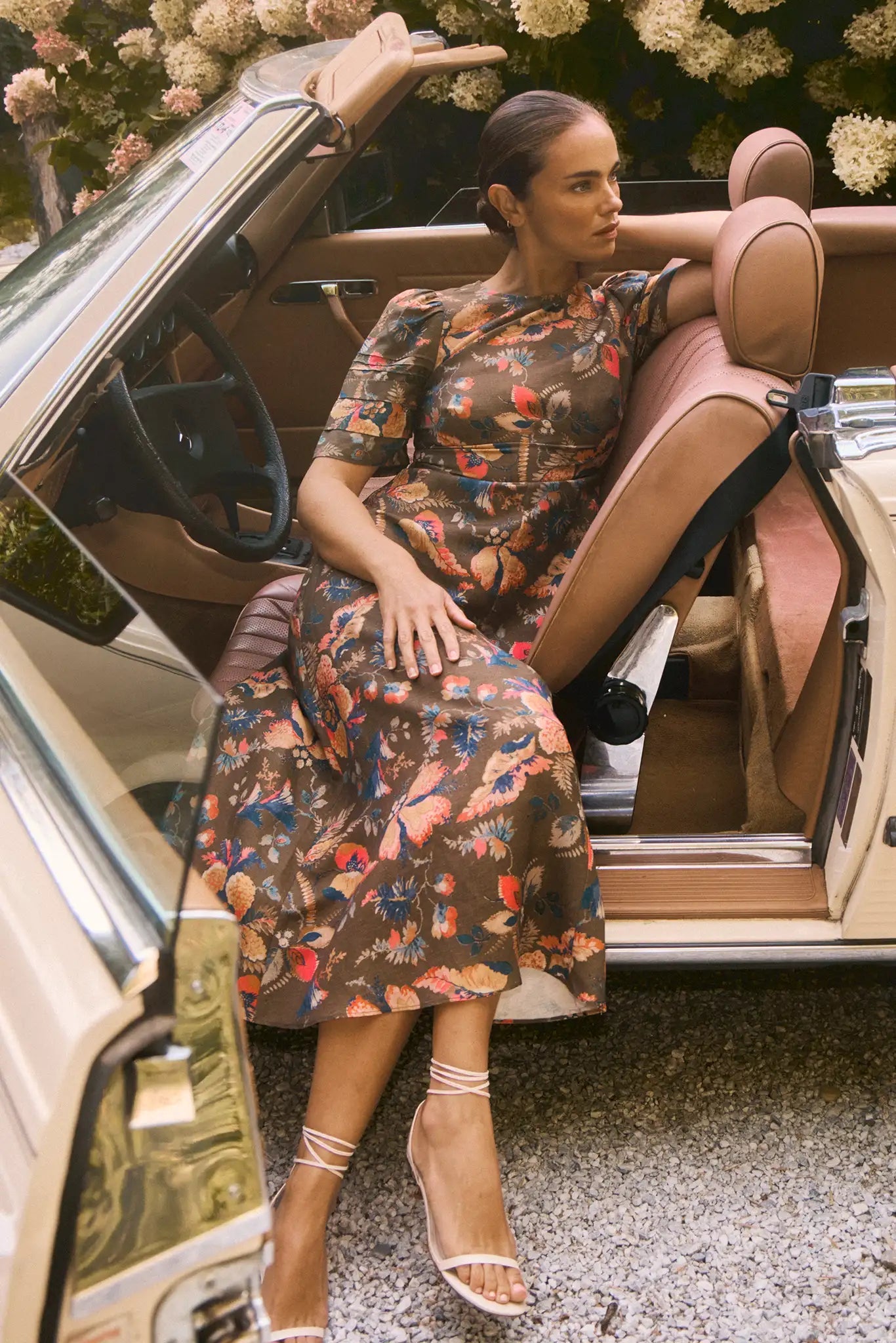 A woman wearing the Casey Dress by Hunter Bell NYC, a universally flattering floral sheath midi, sits in the driver’s seat of a vintage beige convertible with the door open, her arm resting on the back of the seat.