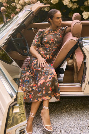 A woman wearing the Casey Dress by Hunter Bell NYC, a universally flattering floral sheath midi, sits in the driver’s seat of a vintage beige convertible with the door open, her arm resting on the back of the seat.