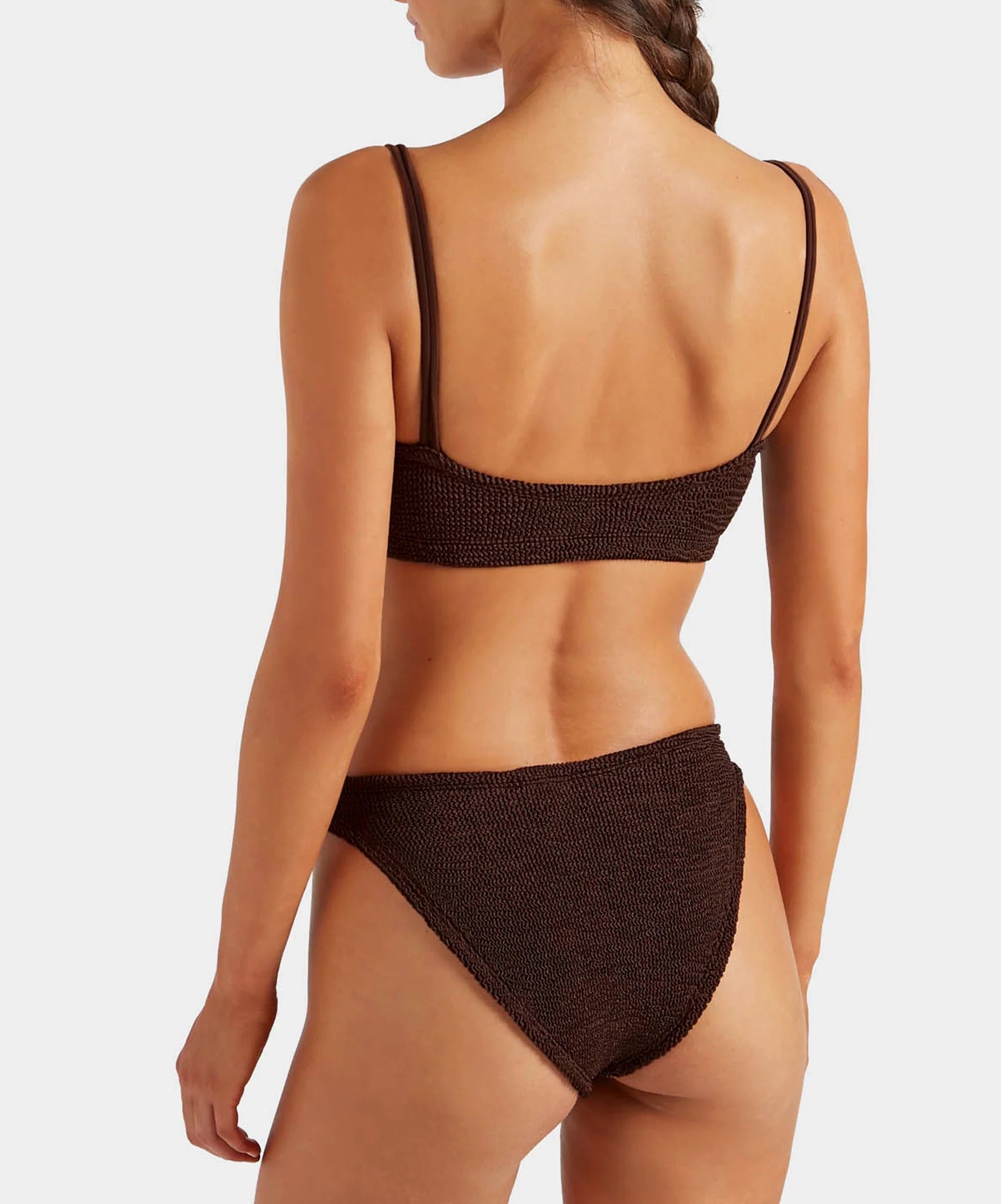 A woman with a braid is shown from the back wearing the Hunza G Cassie Bikini in dark brown, featuring a textured design and classic coverage, set against a plain white background.