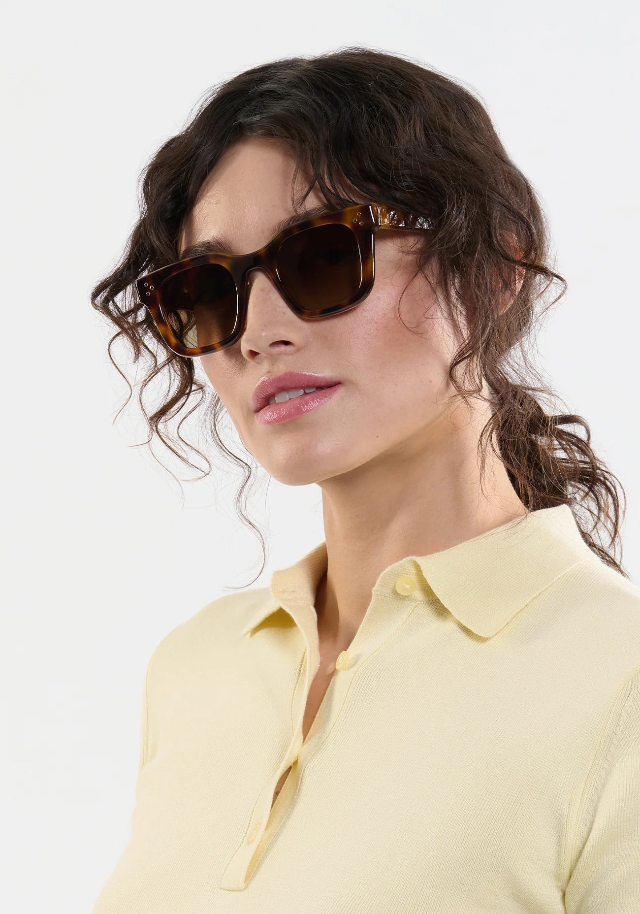Woman with wavy brown hair wears Krewe’s Celeste Polarized Sunglasses, featuring a classic tortoiseshell frame, paired with a light yellow collared shirt against a plain white background.