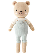 Charlie the Honey Bear by Cuddle + Kind is a 13" plush doll wearing light blue knit overalls and a cream shirt, featuring embroidered facial details and pink cheeks—a sweet companion for family moments.