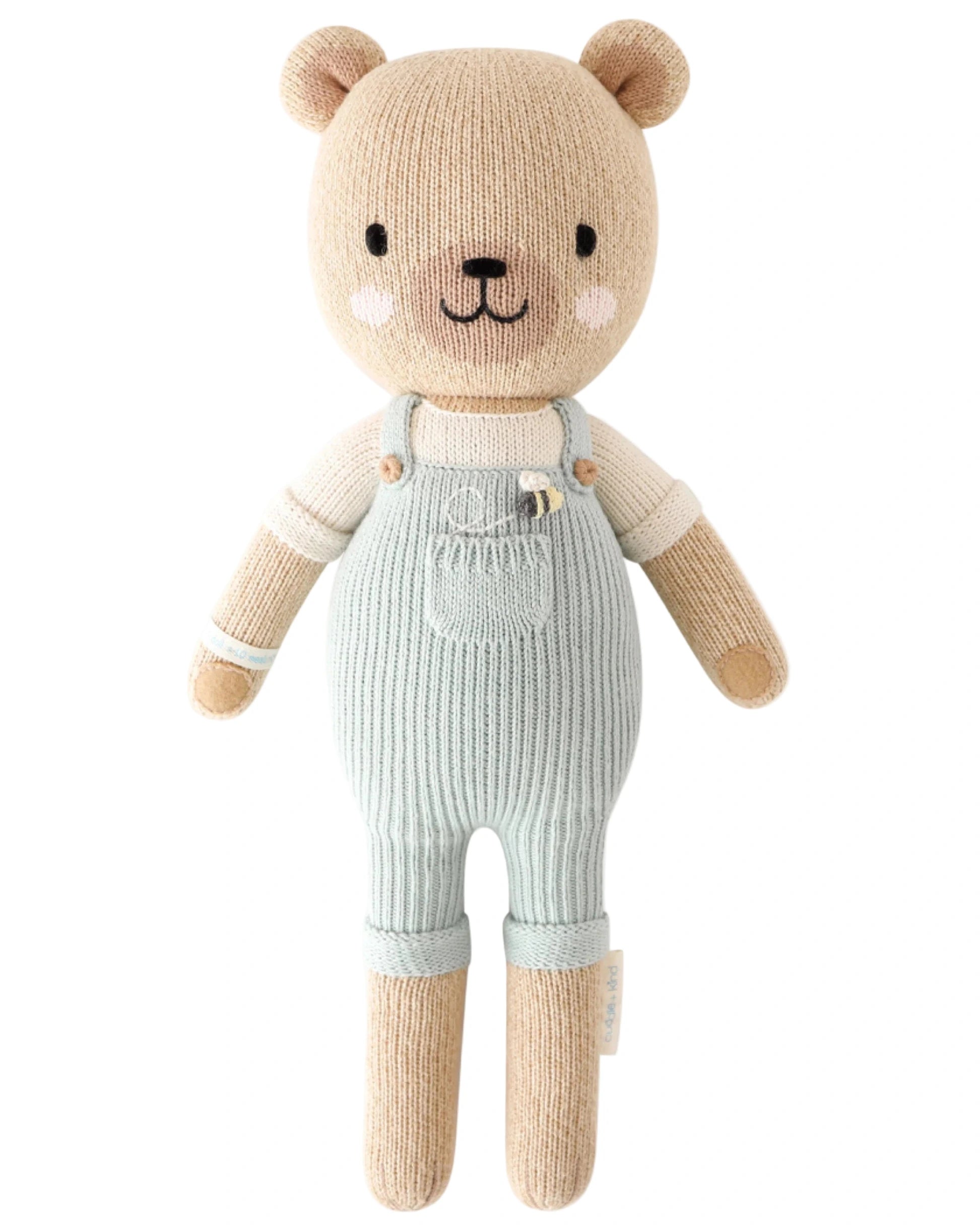 Charlie the Honey Bear by Cuddle + Kind is a 20" knitted teddy in light blue overalls and a cream shirt, with a sweet smile and rosy cheeks—an adorable companion for any family.