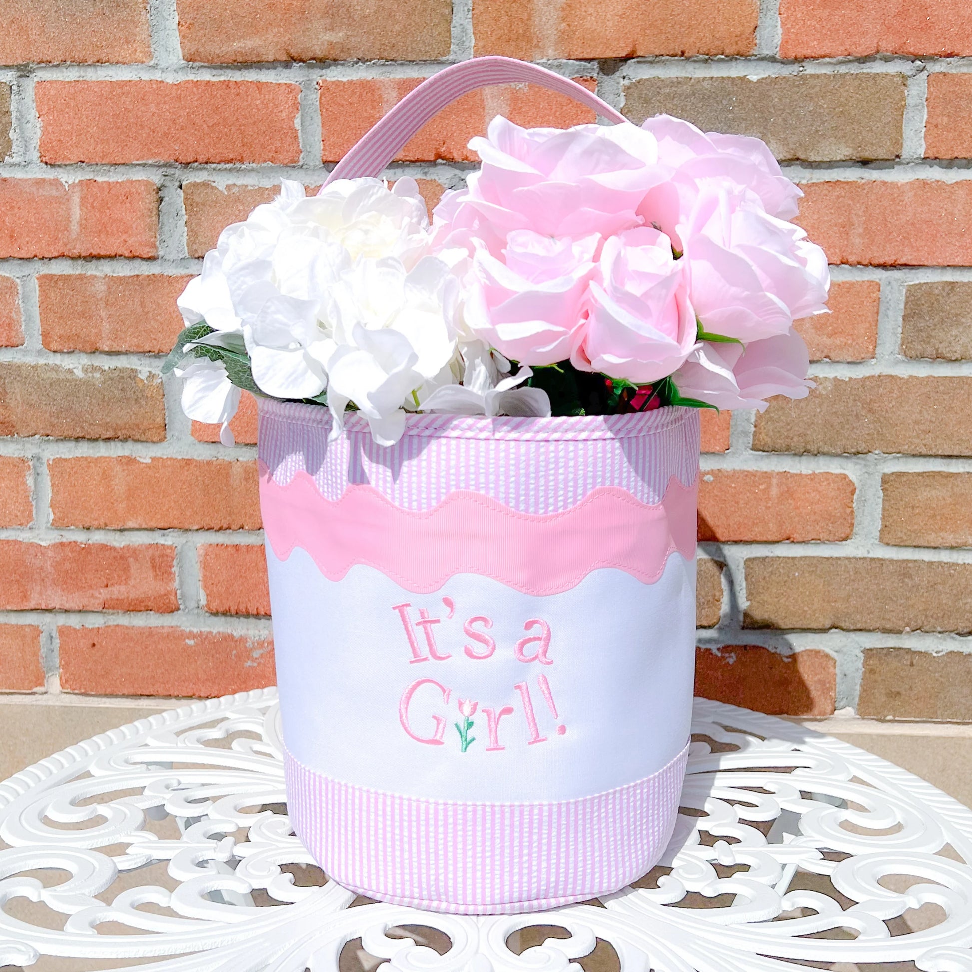The Bits & Bows "It's a Girl Tote," filled with white and pink artificial flowers, sits on a round white table against a brick wall—a delightful gift for new moms.