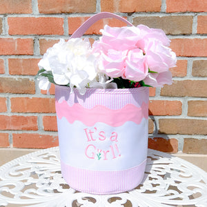 The Bits & Bows "It's a Girl Tote," filled with white and pink artificial flowers, sits on a round white table against a brick wall—a delightful gift for new moms.