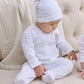 A baby in a Kissy Kissy white and blue Pima cotton onesie and the Cheerful Good Night Printed Hat sits on a light-colored floor by a tufted sofa, looking down with one hand on their leg.