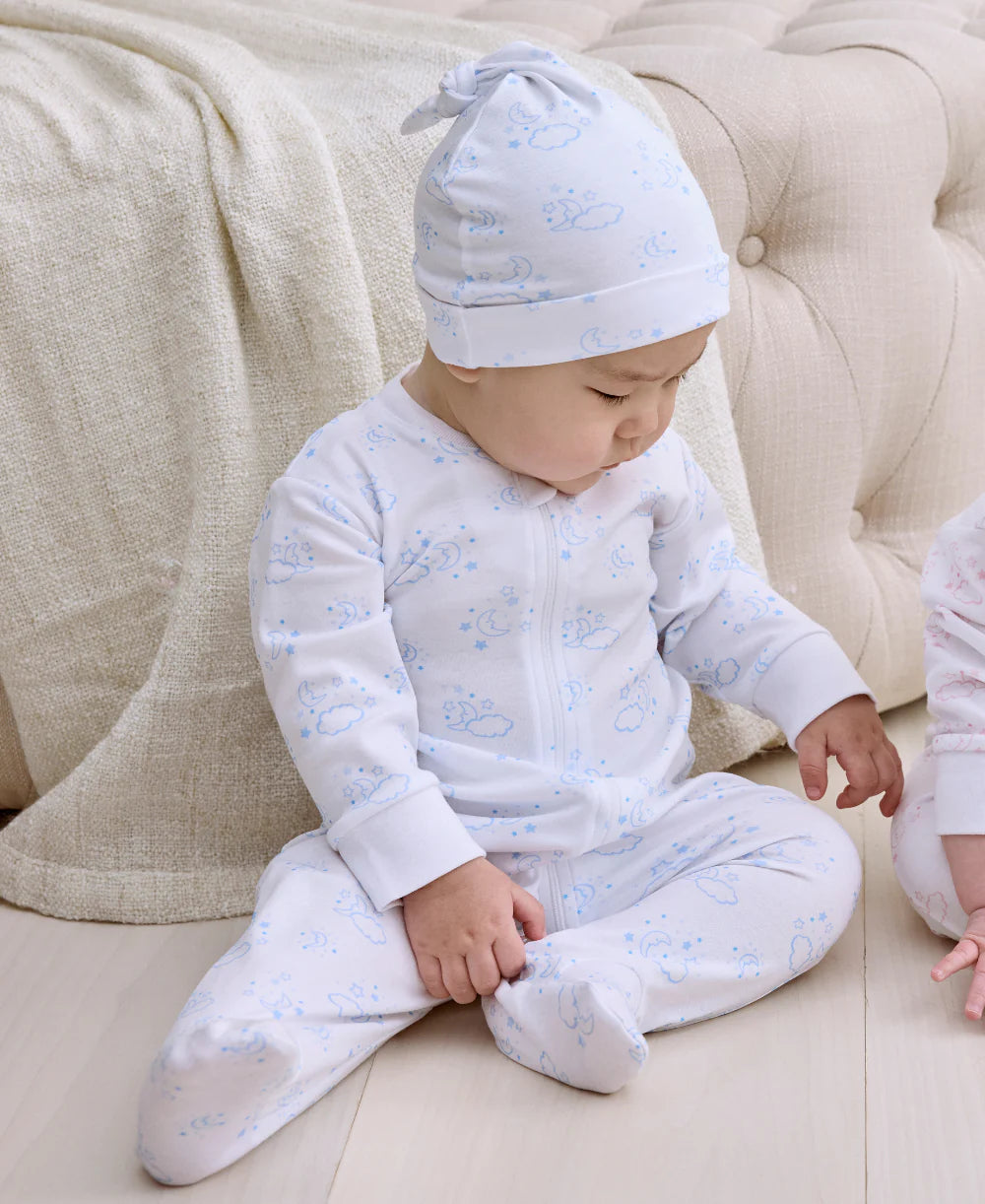 A baby in a Kissy Kissy white and blue Pima cotton onesie and the Cheerful Good Night Printed Hat sits on a light-colored floor by a tufted sofa, looking down with one hand on their leg.