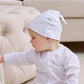 A baby in a Kissy Kissy Cheerful Good Night Printed Hat and pajamas sits on the floor next to a beige tufted couch, reaching toward the furniture.