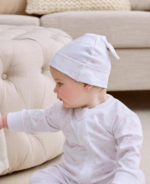 A baby in a Kissy Kissy Cheerful Good Night Printed Hat and pajamas sits on the floor next to a beige tufted couch, reaching toward the furniture.