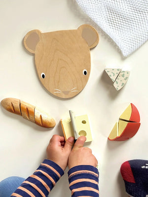 A child's hands play with the Tender Leaf Cheese Chopping Board by Tender Leaf Toys, which features a wooden animal face and a mousey cheese knife on a white surface. This delightful set includes velcro for easy cutting, providing an engaging interactive experience.