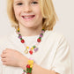 A blonde-haired child smiles in a white shirt, wearing Super Smalls' Make It Fruity Mini Bead Kit necklace and bracelet, adorned with colorful, fruit-inspired charms.