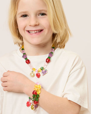 A blonde-haired child smiles in a white shirt, wearing Super Smalls' Make It Fruity Mini Bead Kit necklace and bracelet, adorned with colorful, fruit-inspired charms.