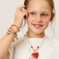 A young girl in a white shirt, smiling and touching her face, wears Super Smalls kids jewelry and shows off her favorite Strawberry Surprise Lipgloss Locket—perfect for beauty play or sharing a fun look.