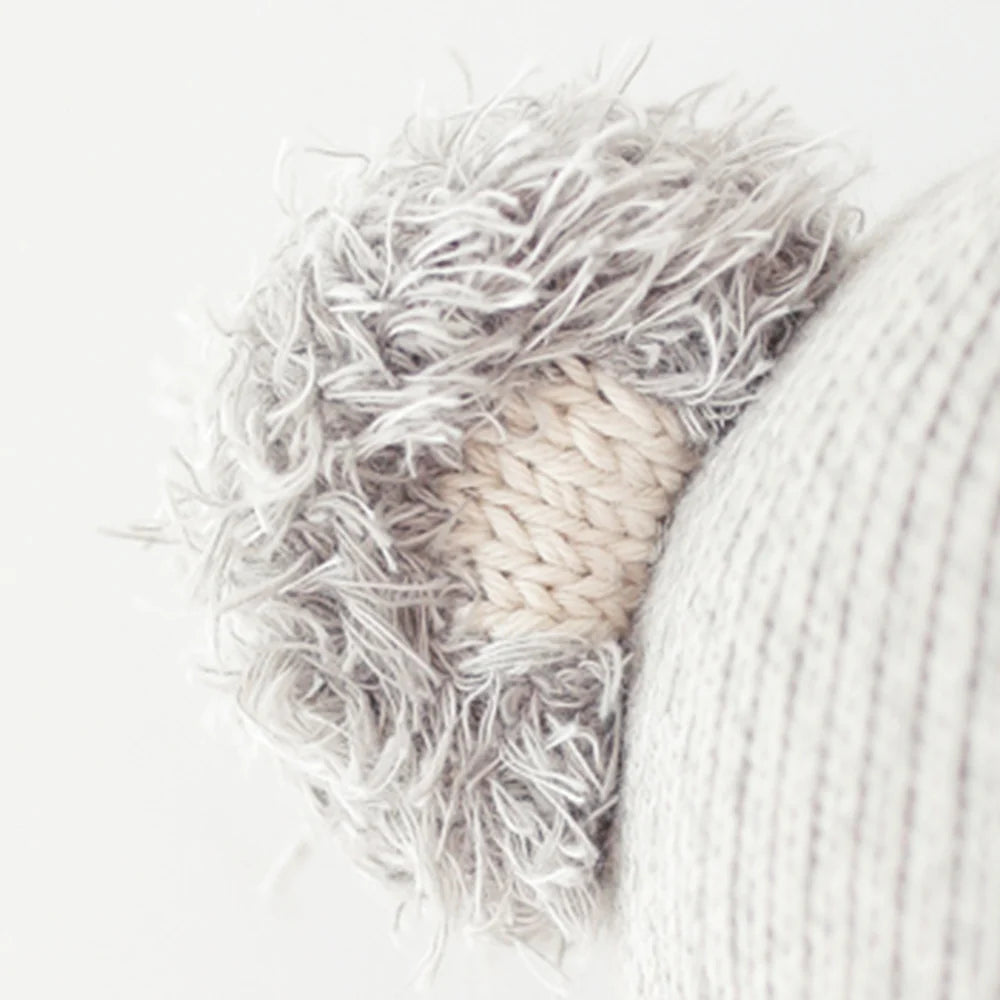 Close-up of a knitted grey and cream ear on Claire the Koala - Regular 20" by Cuddle + Kind, attached to a textured fabric—ideal for snuggling with friends during magical adventures or relaxing in your own cozy space.
