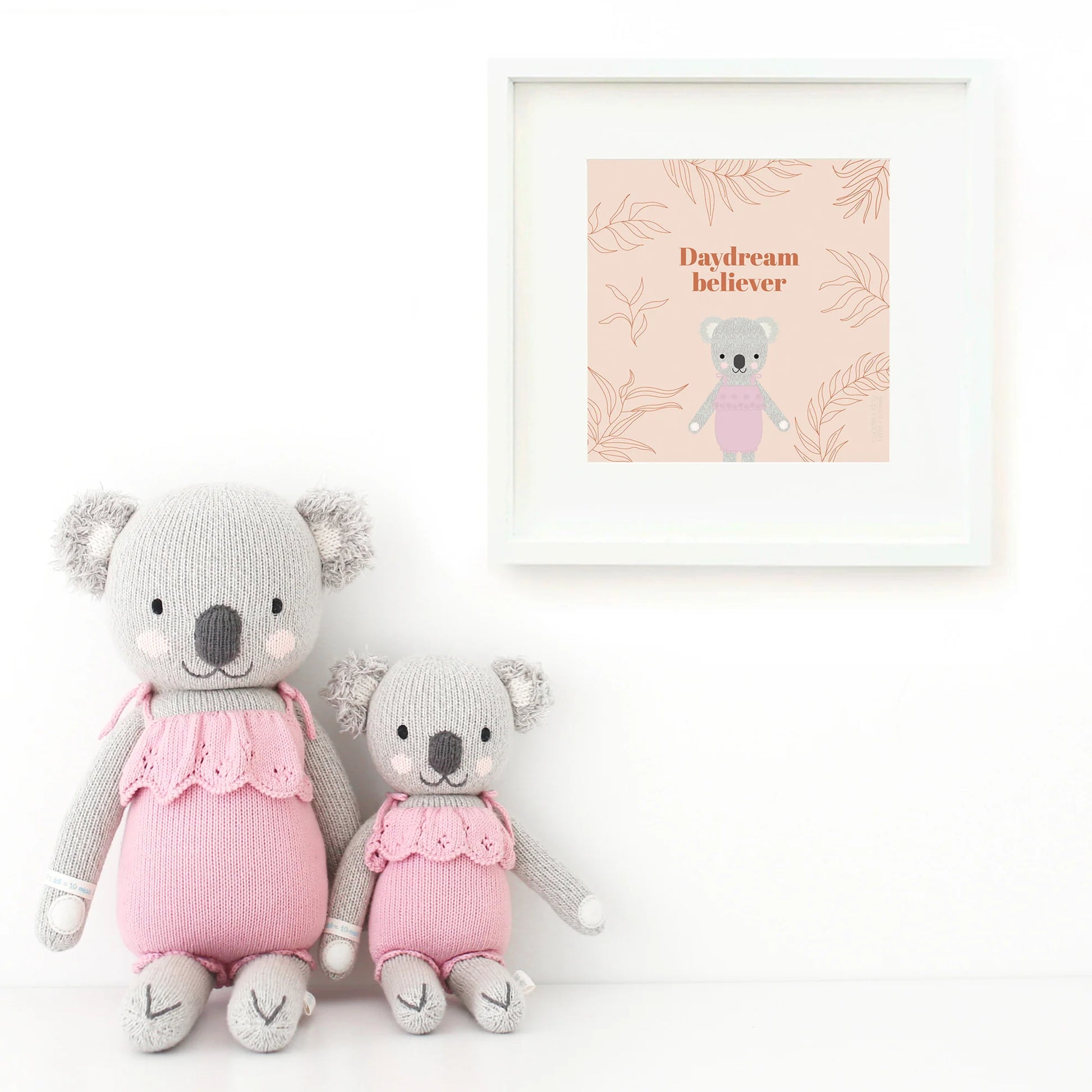 Two Claire the Koala - Regular 20" plush toys from Cuddle + Kind, dressed in pink outfits, sit side by side as best friends beneath a “Daydream believer” print with whimsical koala art and leafy treetops.