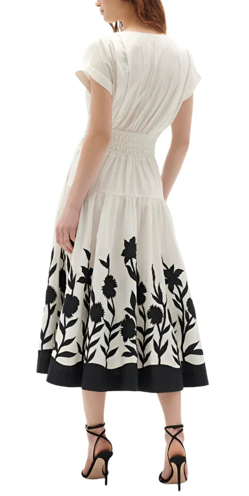 A woman in a Figue Clarette Dress, characterized by its white fabric adorned with black floral patterns and featuring a smocked waist, is seen from behind wearing black strappy heels. Crafted from 100% cotton poplin, this piece beautifully combines elegance with comfort.