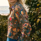 A woman with long blonde hair wears the Clarke Shirt by Hunter Bell NYC, a modern collared top with a floral pattern and jeans, standing outdoors near greenery with the ocean in the background.