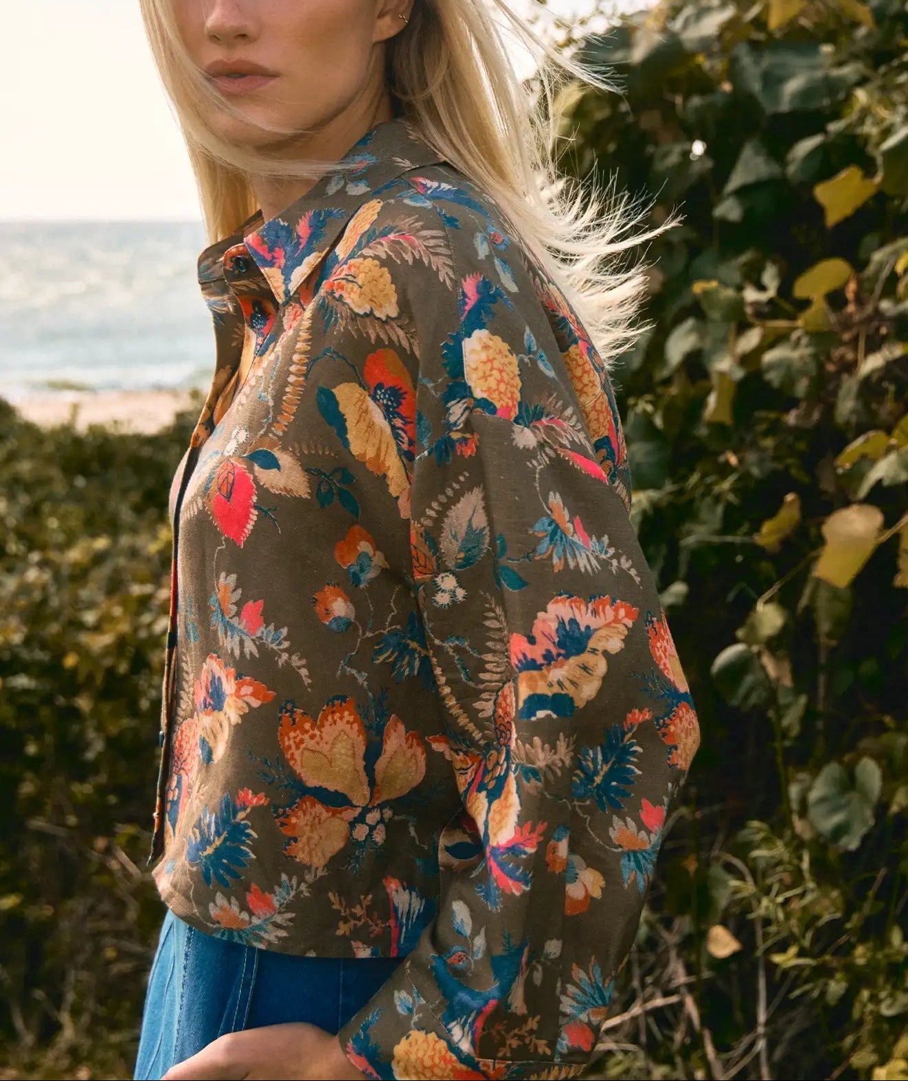 A woman with long blonde hair wears the Clarke Shirt by Hunter Bell NYC, a modern collared top with a floral pattern and jeans, standing outdoors near greenery with the ocean in the background.