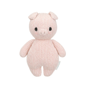 The Piglet - Baby 7" by Cuddle + Kind is a pink, hand-knit stuffed pig made from heirloom-quality Peruvian cotton yarn, featuring black eyes and a small snout. It stands upright against a white background.