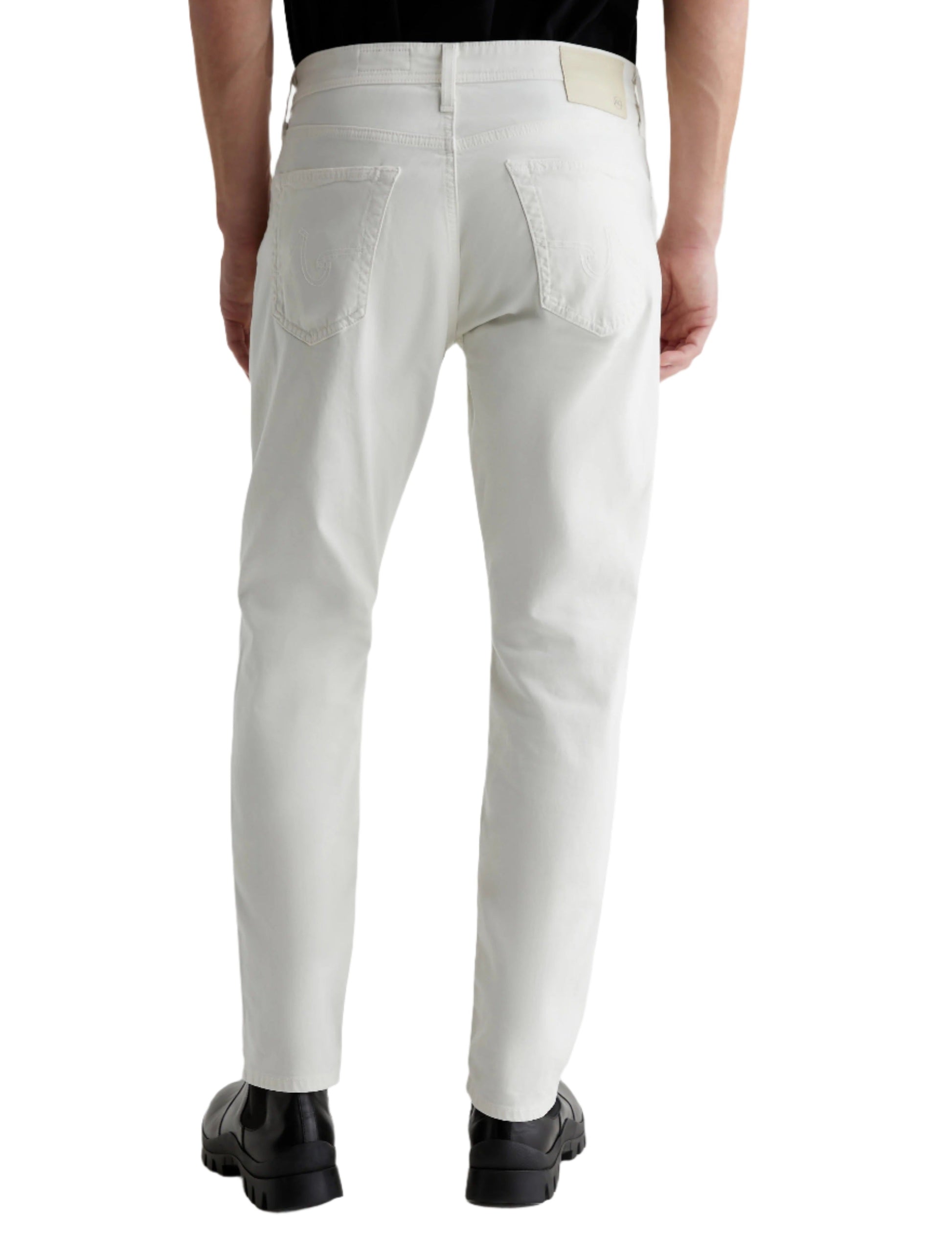 A person wearing AG Jeans Everett Sueded in white, a black shirt, and black shoes is shown from the back against a plain background. The sueded stretch sateen fabric of the jeans offers a comfortable mid-rise waist for ease of movement.