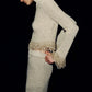 A model wears a shimmering long-sleeved knit top paired with a metallic bouclé Zeni Midi Skirt, featuring dangling pearl fringe trim at the cuffs and hem.