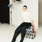 Wearing the Aaliyah Knit Turtleneck by Simkhai in ivory, a person pairs it with black textured pants while seated on stacked colorful trunks in a studio set against a white backdrop with lights.
