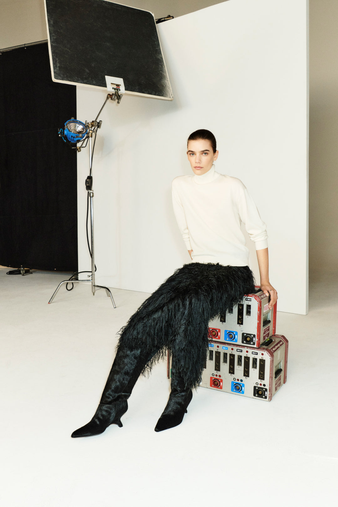 Wearing the Aaliyah Knit Turtleneck by Simkhai in ivory, a person pairs it with black textured pants while seated on stacked colorful trunks in a studio set against a white backdrop with lights.