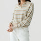 A woman wearing the Guest in Residence Collegiate Stripe Polo—a beige and light blue striped cashmere sweater with a collar—paired with white pants, stands against a plain light background.