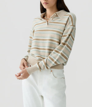 A woman wearing the Guest in Residence Collegiate Stripe Polo—a beige and light blue striped cashmere sweater with a collar—paired with white pants, stands against a plain light background.