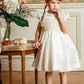 A young girl in an Antoinette Girls' Colombe Gold Silk Ceremony Dress and tiara stands beside a table with flowers, looking thoughtful.