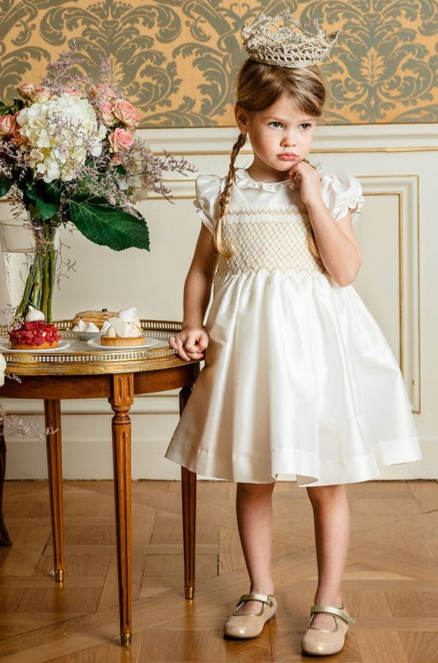 A young girl in an Antoinette Girls' Colombe Gold Silk Ceremony Dress and tiara stands beside a table with flowers, looking thoughtful.