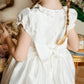 Young girl with braided hair, seen from behind, wearing a white Antoinette Girls' Colombe Gold Silk Ceremony Dress in a floral decorated room.