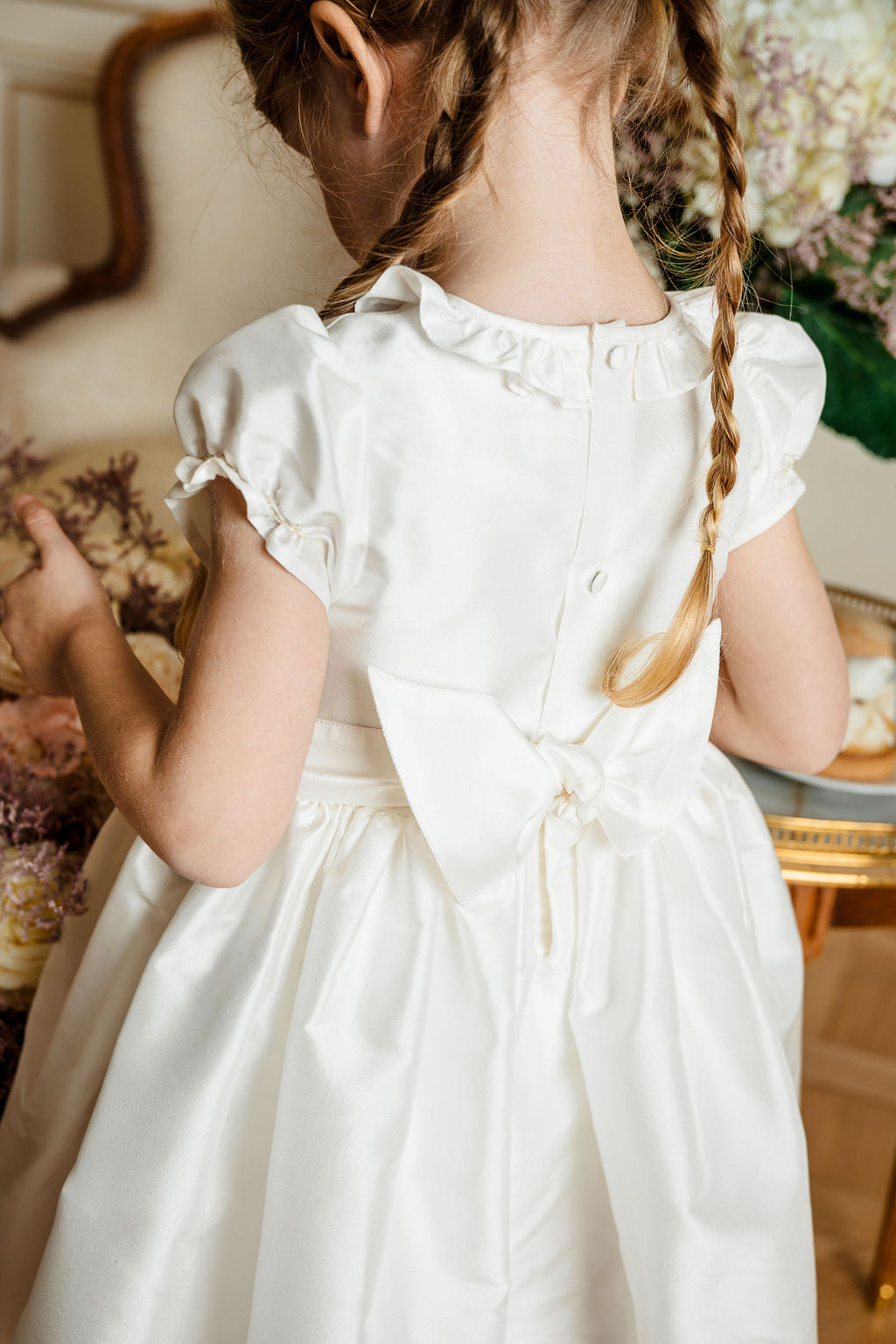 Young girl with braided hair, seen from behind, wearing a white Antoinette Girls' Colombe Gold Silk Ceremony Dress in a floral decorated room.