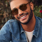 A man wearing Krewe Colton Tortuga Silver sunglasses, a denim jacket, and a light sweater smiles outdoors with greenery in the background.