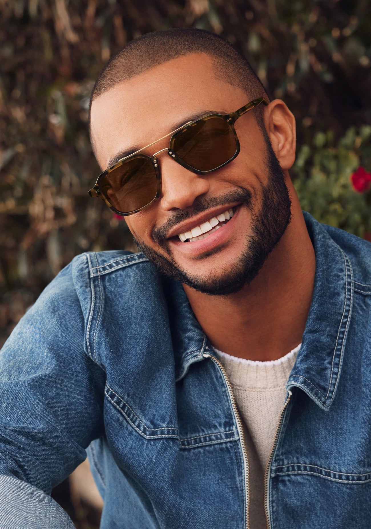 A man wearing Krewe Colton Tortuga Silver sunglasses, a denim jacket, and a light sweater smiles outdoors with greenery in the background.