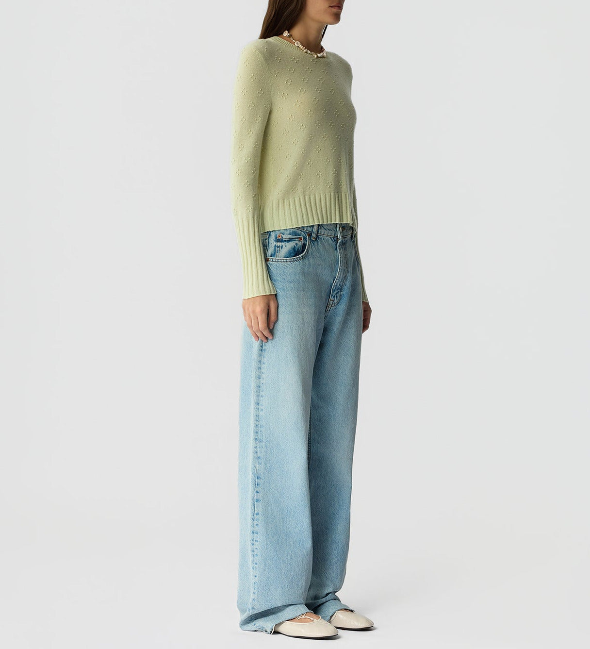 A person stands against a plain background wearing the Compass Shrunken Crew cashmere sweater by Guest in Residence, wide-leg blue jeans, and white shoes.