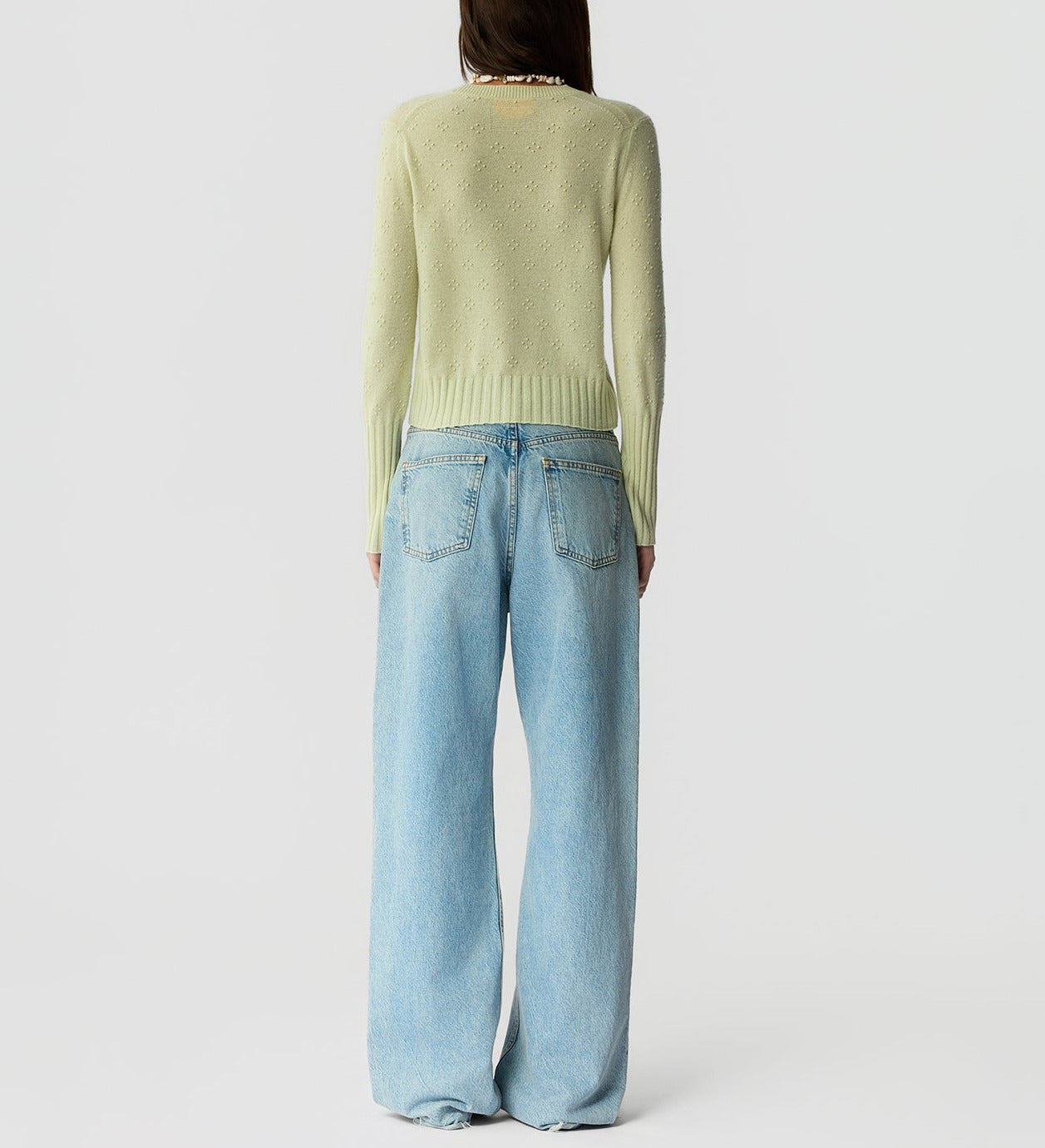 A person stands with their back to the camera, wearing the Guest in Residence Compass Shrunken Crew sweater in light green and loose light blue jeans against a plain background.