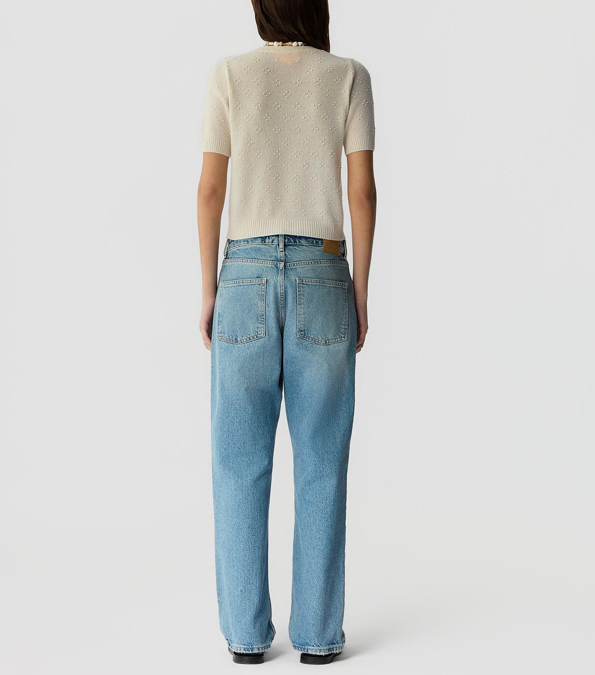 A person stands facing away, wearing the Compass Sweater Tee by Guest in Residence— a women's lightweight cashmere crew neck short-sleeve sweater— with loose-fitting light blue jeans against a plain white background.