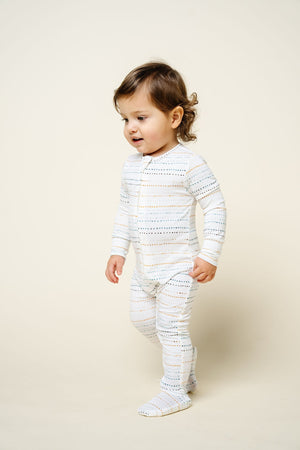 A toddler stands and walks on a light background, dressed in Lou Lou & Company’s Baby Conrad Zipper Footie—silky-soft, white sleepwear featuring small, colorful dotted lines for cozy comfort.