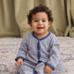 A baby with curly hair sits on a patterned rug, smiling at the camera while wearing the Kissy Kissy Baby Boys’ Construction Demo Striped Footie—a blue and white striped wardrobe staple. A green curtain hangs in the background.
