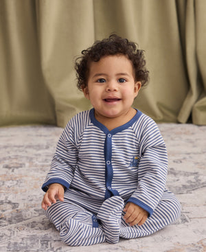 A baby with curly hair sits on a patterned rug, smiling at the camera while wearing the Kissy Kissy Baby Boys’ Construction Demo Striped Footie—a blue and white striped wardrobe staple. A green curtain hangs in the background.