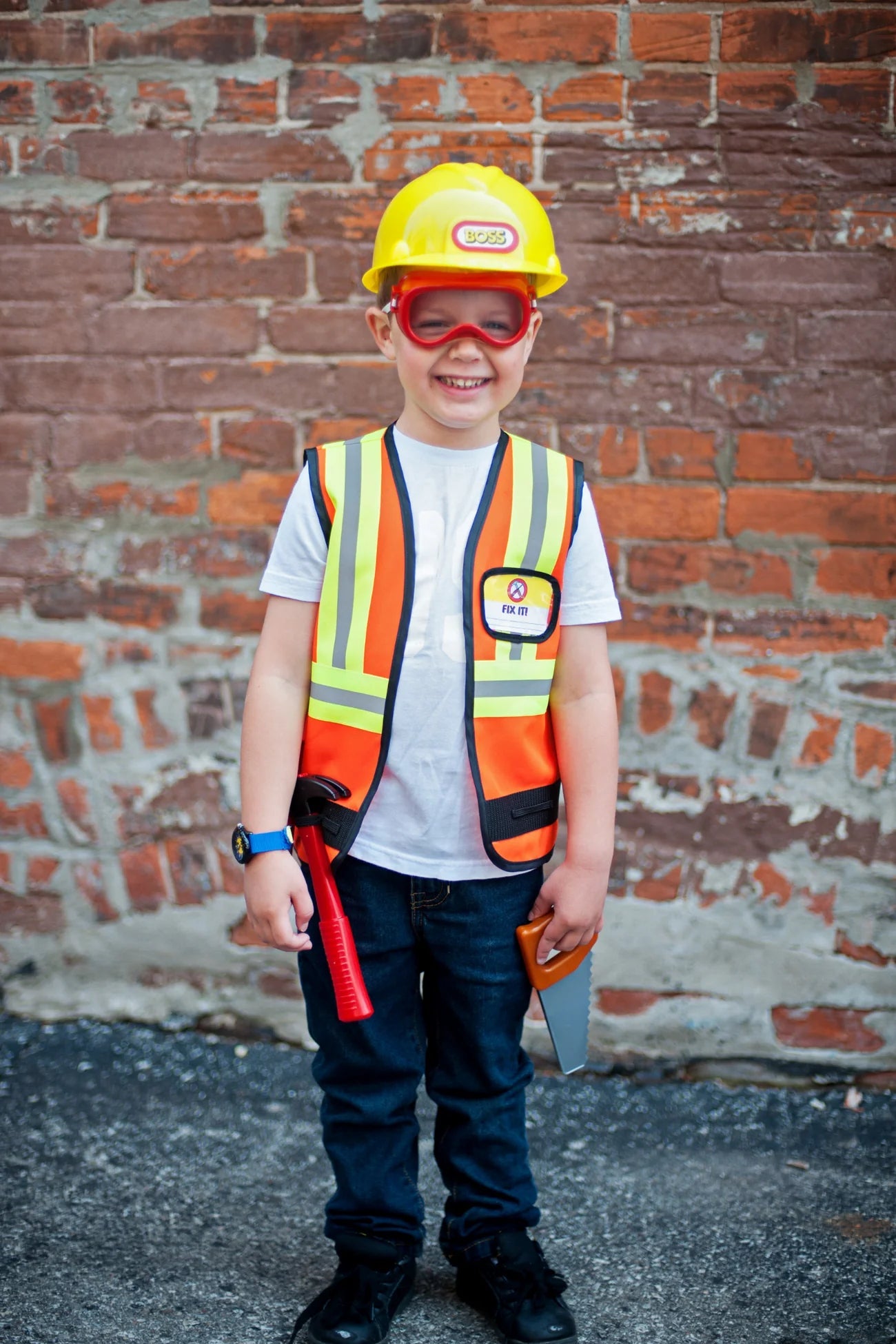 A child beams in front of a brick wall, proudly holding a toy tool while wearing the Great Pretenders Construction Worker Set—complete with helmet, safety goggles, and vest—for fun dress up play.